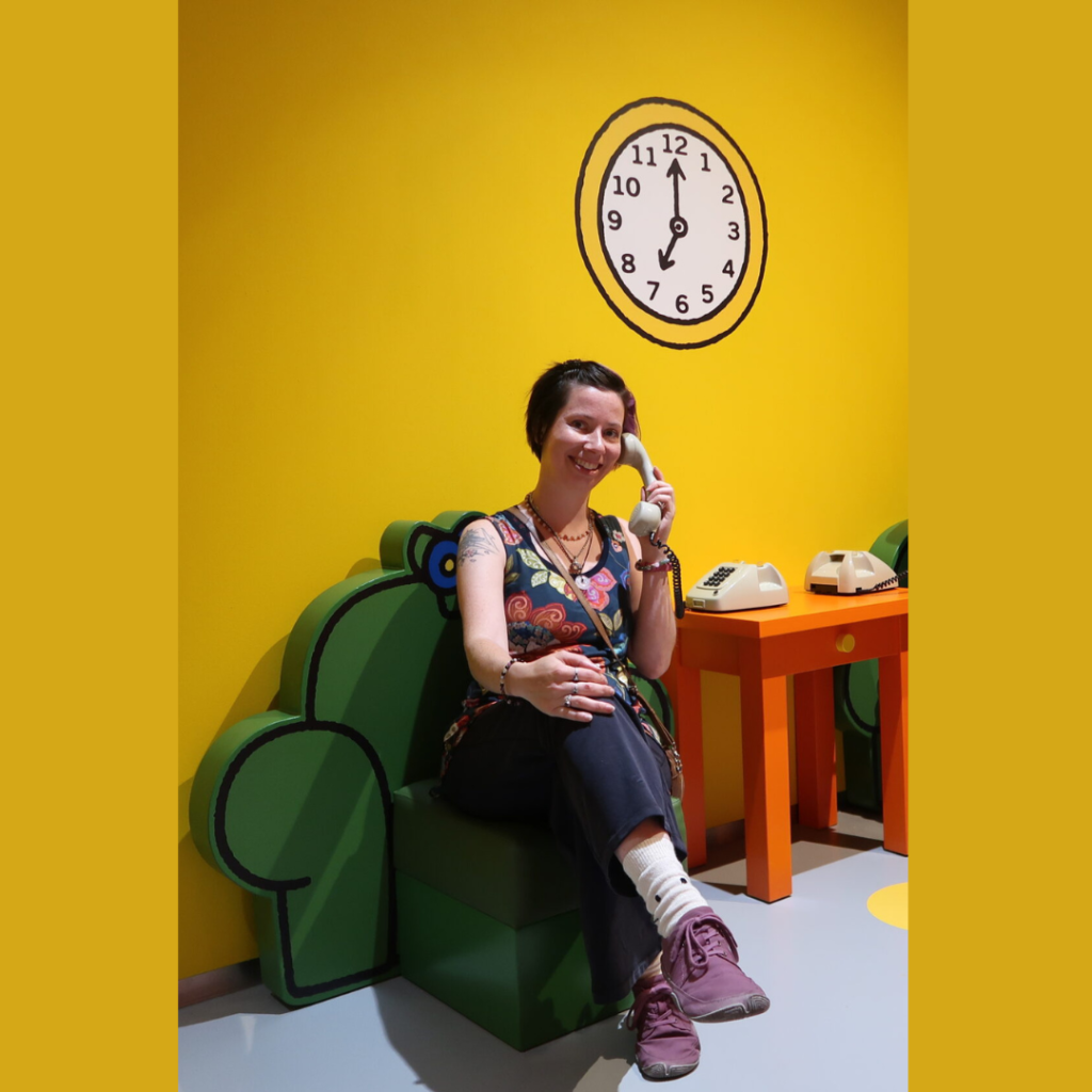 Leyla op de foto in het Nijntje museum. Zittend op een groene cartoon stoel, met een draai telefoon in haar hand.